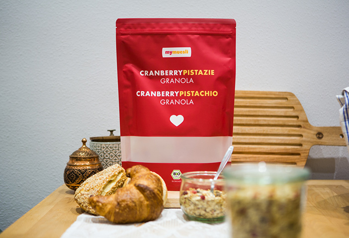 Eine rote Müsli Verpackung steht in der Mitte von einem Croissant und einem Müsli in einem Becher