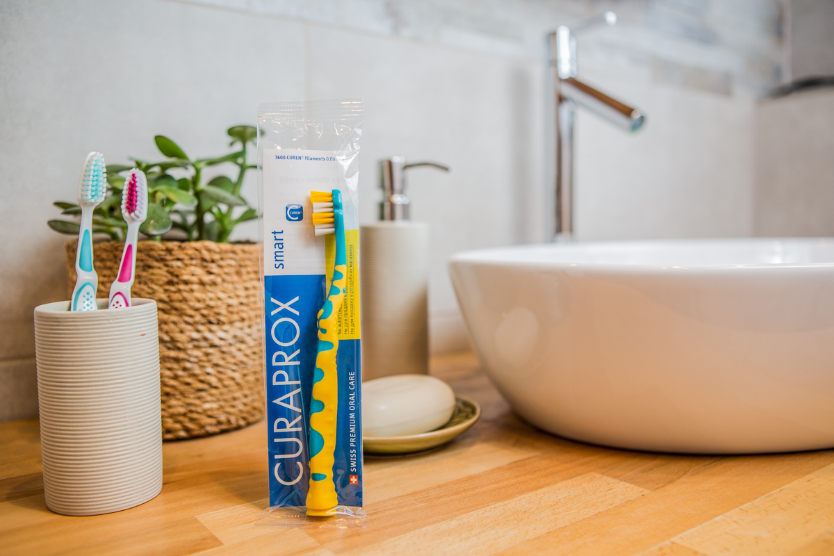 Mehrer Zahnbürsten, eine Seife, ein Zahnputzbecher und eine Pflanze liegen an einem Waschbecken.