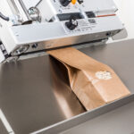 Durchlaufsiegelgerät MSC beim Siegeln eines Verbundmaterials mit Tischstativ und neigbarer Beutelauflage.