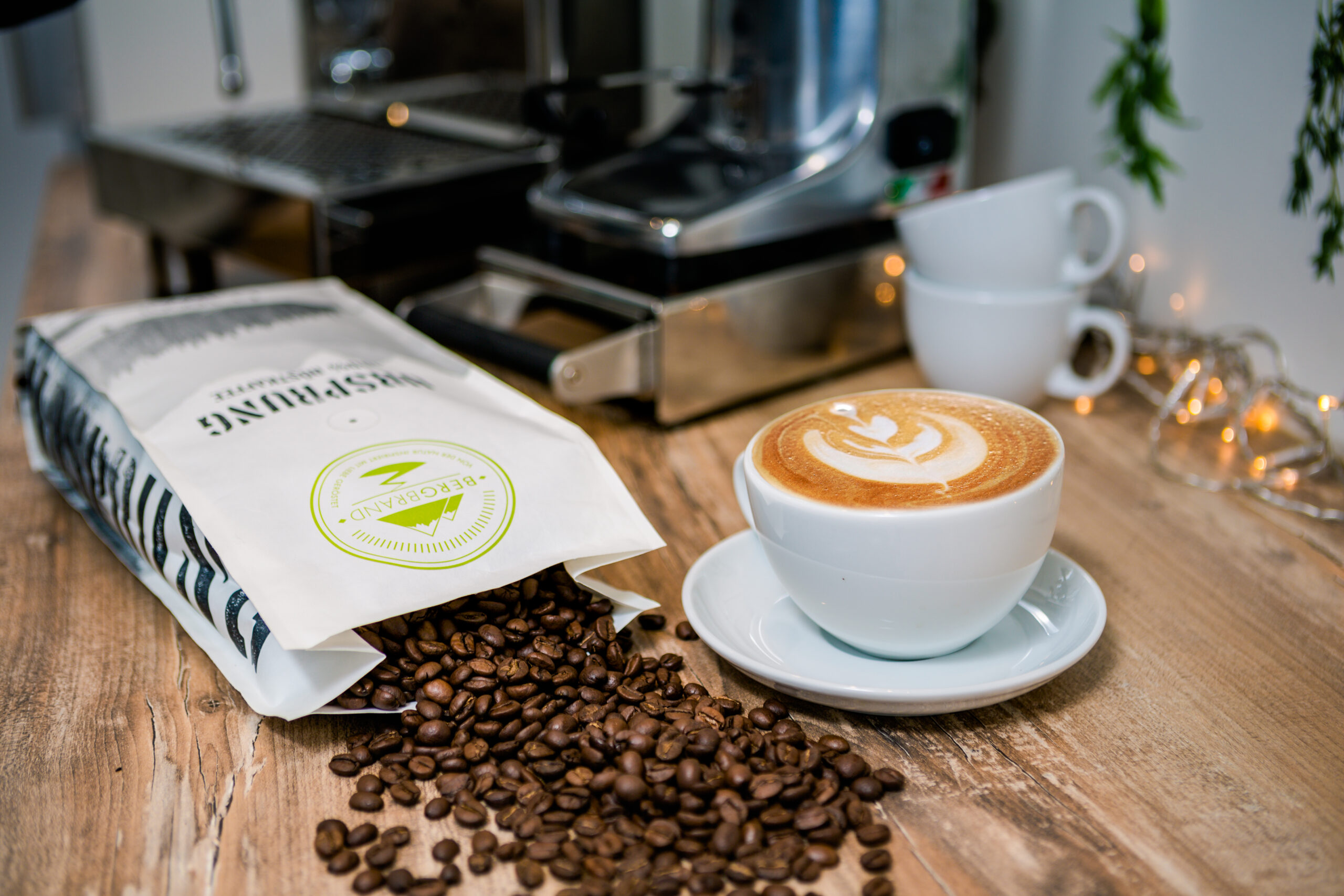 Ein Café und eine geöffnete Kaffeeverpackung liegen auf einem Tisch. Im Hintergrund steht eine Kaffeemaschine und Kaffeetassen