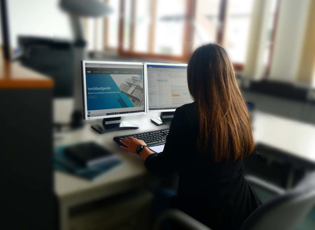 Eine Frau mit braunen Haaren sitzt vor einem Computer auf einem Bürostuhl. Sie arbeitet gerade an dem Computer.