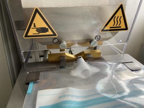 Auf dem Bild werden Spouts in eine Folie eingesiegelt. Links und rechts sind Warnsymbole zu sehen.