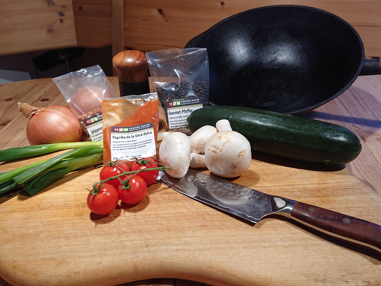 Verschiedene Gewürze und Gemüse liegen mit einem Messer und einer Pfanne auf einem Tisch.