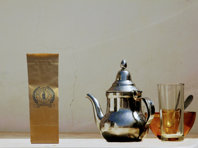 Ein Teebeutel, eine Teekanne, Glas und dahinter eine Schüssel stehen auf einem Tisch.