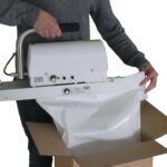 Mit einem mobilen Durchlaufschweißgerät wird ein weißer Beutel in einem Karton verschweißt. Der Anwender hält das Gerät am Griff und mit der anderen Hand den Beutel.